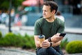 Cheerful young man using mobile phone while sitting at the city park, drinking coffee Royalty Free Stock Photo