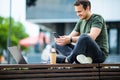 Cheerful young man using mobile phone while sitting at the city park, drinking coffee Royalty Free Stock Photo