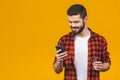 Cheerful young man wearing plaid shirt standing isolated over yellow background, holding mobile phone, showing plastic credit card Royalty Free Stock Photo