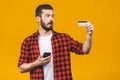 Cheerful young man wearing plaid shirt standing isolated over yellow background, holding mobile phone, showing plastic credit card Royalty Free Stock Photo