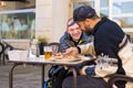 Cheerful young man in a wheelchair with his companion enjoying their meal outdoors on a sunny day