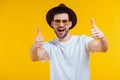 cheerful young man in white t-shirt, hat and sunglasses showing thumbs up and smiling at camera