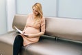 Cheerful young pregnant woman is sitting in hallway of hospital waiting to see gynecologist. Royalty Free Stock Photo