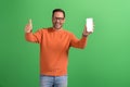 Cheerful young salesman showing thumbs up sign and smart phone's screen on isolated green background