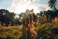 Cheerful young stylish woman holding a bunch of banans over the head on the feild at sunset