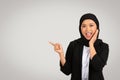 A cheerful young woman in a black hijab and blazer is pointing to the side with one hand while touching her face Royalty Free Stock Photo