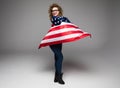 Cheerful young woman in casual clothes and glasses is covered in the American flag and smiling on gray background Royalty Free Stock Photo