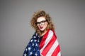 Cheerful young woman in casual clothes and glasses is covered in the American flag and smiling on gray background Royalty Free Stock Photo