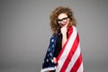 Cheerful young woman in casual clothes and glasses is covered in the American flag and smiling on gray background Royalty Free Stock Photo