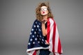 Cheerful young woman in casual clothes and glasses is covered in the American flag and smiling on gray background Royalty Free Stock Photo