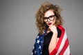 Cheerful young woman in casual clothes and glasses is covered in the American flag and smiling on gray background Royalty Free Stock Photo