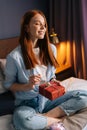 Cheerful young woman opening gift box with present with closed eyes at cozy living room near laptop. Royalty Free Stock Photo