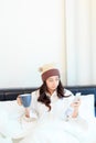 Cheerful young woman sitting in bed texting on her smartphone and drinking coffee at home in bedroom Royalty Free Stock Photo