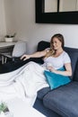 Cheerful young woman sitting on a couch at home while using a remote tv controller Royalty Free Stock Photo