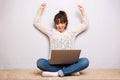 Cheerful young woman sitting on floor with laptop and arms raised Royalty Free Stock Photo