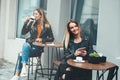 Cheerful young woman sitting outdoor drinking coffee smiling and holding smartphone in hand with beautiful fashionable girl on bac Royalty Free Stock Photo