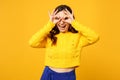 Cheerful young woman in sweater blue trousers hold hands near eyes imitating glasses or binoculars isolated on yellow Royalty Free Stock Photo