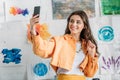 Young woman taking selfie while standing near white wall with colorful paintings