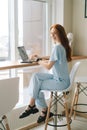 Cheerful young woman wearing casual clothing typing on laptop computer sitting at table by window in cafe, looking at Royalty Free Stock Photo