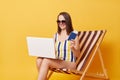 Cheerful young woman wearing swimsuit sit on chair booking tickets online holding passport and laptop isolated over yellow Royalty Free Stock Photo