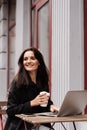 Cheerful young woman is working online on laptop and sitting on summer terrace in cafe. Happy excited girl with cup of Royalty Free Stock Photo