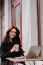 Cheerful young woman is working online on laptop and sitting on summer terrace in cafe. Happy excited girl with cup of Royalty Free Stock Photo