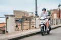 Cheerfully smiling man in helmet and sunglasses fast riding the moto scooter on the Sicilian old town streets in the Forza d`Agro