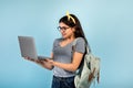 Cheery Indian teen girl with backpack and laptop pc having online lesson on blue studio background Royalty Free Stock Photo