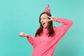 Cheery woman in knitted pink sweater birthday hat keeping mouth wide open showing victory sign, hold in hand cake with