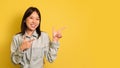 Cheery young Asian lady pointing at copy space, smiling, offering place for your ad design over yellow background