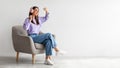 Cheery young lady in headphones listeing to music and dancing while sitting in armchair against white studio wall