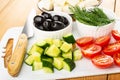 Cheese in brown saucer, bowls with dill, black olives, pieces of cucumbers, tomato, knife on cutting board on table Royalty Free Stock Photo