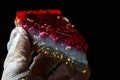 Cheese cake with red strawberry jelly on top held in left hand in hygienic latex glove, dark background