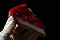 Cheese cake with red strawberry jelly on top held in left hand in hygienic latex glove, dark background