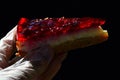 Cheese cake with red strawberry jelly on top held in left hand in hygienic latex glove, dark background