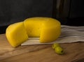 Cheese wheel with a cut piece on a napkin. In the background, green olives in a plate and part of a bottle of wine