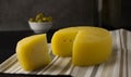 Cheese wheel with a cut piece on a napkin. In the background, green olives in a plate and part of a bottle of wine