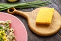 Cheese on a wooden board next to green onions on the table and a fragment of a plate with pasta in a creamy sauce