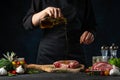 The chef in black apron pours oil on the fresh beef or pork steak on the wooden chopping board. Frozen motion. Backstage of