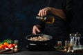 The chef in black apron pours oil into the pan with frying meat on dark blue background. Professional kitchen table with spices, Royalty Free Stock Photo