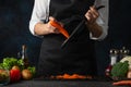 The chef in black apron shredding carrots with grater on black chopped board at professional restaurant kitchen on dark blue