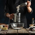 The chef in black apron sifts flour into glass bowl for preparing dough. Backstage of cooking waffle on rustic wooden table with Royalty Free Stock Photo
