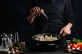 The chef in black uniform adds the pepper to the frying chicken in the wog pan isolated on the dark background. Restaurant`s dish