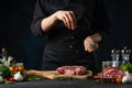 The chef in black uniform pours rosemary on steak on the wooden board on dark blue background. Preparing raw beef or pork.
