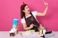 Chef cook confectioner or baker in brown apron, white t-shirt, red hairband, making cake at table, doing selfie on mobile phone,