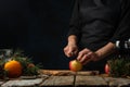 Chef cuts with knife apple on wooden chopped board for preparing mulled wine on rustic wooden table with festive composition