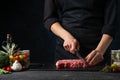 The chef cuts with knife fresh marbled beef on the black board on dark blue background. Backstage of preparing rib-eye steak. Food Royalty Free Stock Photo