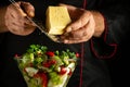 The chef grates cheese into a vitamin salad for lunch. Concept of cooking vegetarian food in a bowl on a black background Royalty Free Stock Photo