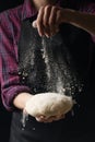 The chef keeps the dough and pours flour on a dark background. The concept of nutrition.