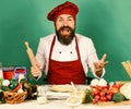 Chef with pasta, vegetables and dough on table. Cook with excited face in burgundy uniform holds rolling pin Royalty Free Stock Photo